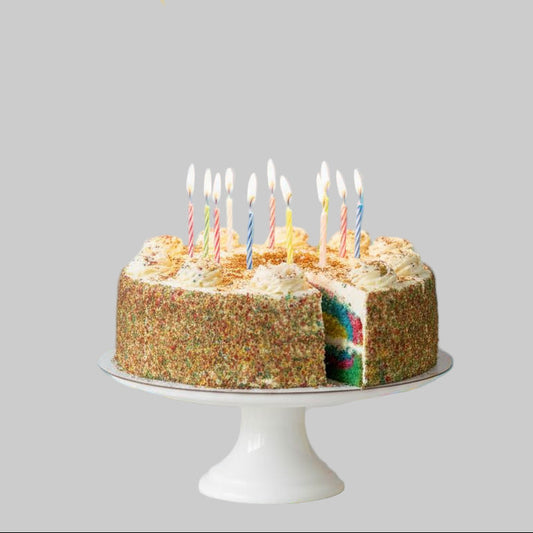 Birthday rainbow cake with lit candles on a white stand against a light gray background.